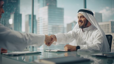 Smiling Emirati Businessman in White Traditional Outfit Sitting in Office with Business Partners. Shake Hands on Successful Investment. Saudi, Emirati, Arab Businessman Concept.
