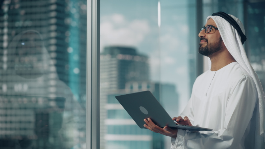 Successful Muslim Businessman in Traditional White Kandura Standing in His Modern Office, Using Laptop Computer Next to Window with Skyscrapers. Successful Saudi, Emirati, Arab Businessman Concept. Royalty-Free Stock Footage #1082792137