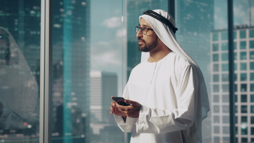 Successful Muslim Businessman in Traditional White Outfit Standing in His Modern Office, Using Smartphone Next to Window with Skyscrapers. Successful Saudi, Emirati, Arab Businessman Concept. Royalty-Free Stock Footage #1082792140