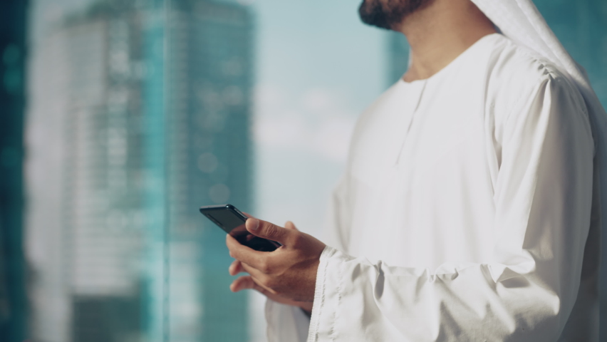 Authentic Muslim Businessman in Traditional White Kandura Standing in His Modern Office, Using Smartphone Next to Window with Skyscrapers. Successful Saudi, Emirati, Arab Businessman Concept. Royalty-Free Stock Footage #1082792146