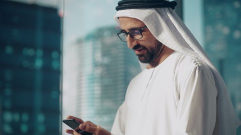 Authentic Muslim Businessman in Traditional White Kandura Standing in His Modern Office, Using Smartphone Next to Window with Skyscrapers. Successful Saudi, Emirati, Arab Businessman Concept.