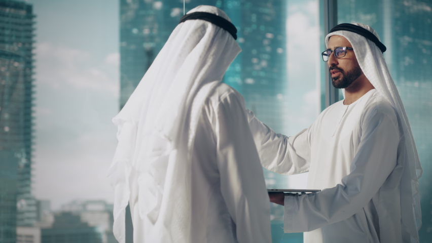 Two Successful Muslim Businessmen in White Traditional Outfit Standing in Office Meeting, Negotiating and Talking About Financial Opportunities, Using Laptop. Saudi, Emirati, Arab Businessman Concept. Royalty-Free Stock Footage #1082792158
