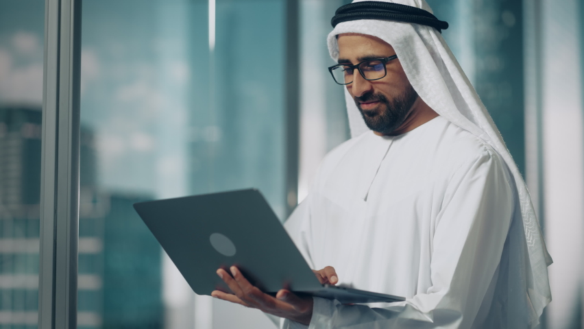 Successful Muslim Businessman in Traditional White Outfit Standing in His Modern Office, Using Laptop Computer Next to Window with Skyscrapers. Successful Saudi, Emirati, Arab Businessman Concept. Royalty-Free Stock Footage #1082792167