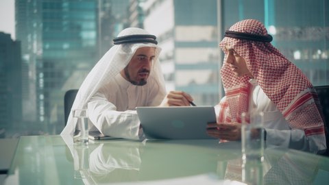 Two Successful Emirati Businessmen in White Traditional Outfit Sitting in Office Meeting, Negotiating and Talking About Financial Opportunities, Using Laptop. Saudi, Emirati, Arab Businessman Concept.