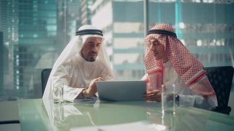 Two Successful Emirati Businessmen in White Traditional Outfit Sitting in Office Meeting Negotiating and Talking About Investment Opportunities, Using Laptop. Saudi, Emirati, Arab Businessman Concept.
