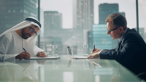 Two Successful Multicultural Businessmen Sitting in Office and Signing Contract. Arab Business Partner Shake Hands with Investor on Lucrative Financial Deal. Saudi, Emirati, Arab Businessman Concept.