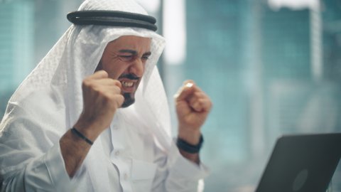 Arab Businessman in White Traditional Outfit Sitting in Office and Working on Laptop Computer. Business Manager Celebrates Successful Investment Deal. Saudi, Emirati, Arab Businessman Concept.