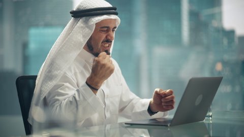 Arab Businessman in White Traditional Outfit Sitting in Office and Working on Laptop Computer. Business Manager Celebrates Successful Investment Deal. Saudi, Emirati, Arab Businessman Concept.