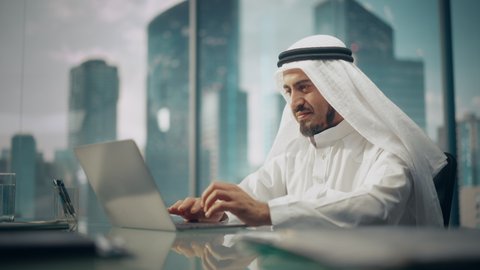 Successful Arab Businessman in White Traditional Kandura Sitting in Office and Working on Laptop Computer. Business Manager Planning Investments. Saudi, Emirati, Arab Businessman Concept.