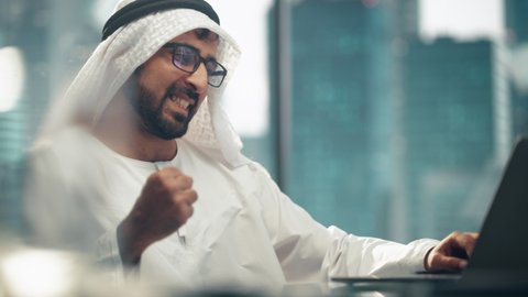 Successful Arab Businessman in Glasses and White Traditional Outfit Sitting in Office and Working on Laptop. Business Manager Planning Corporate Strategy. Saudi, Emirati, Arab Businessman Concept.