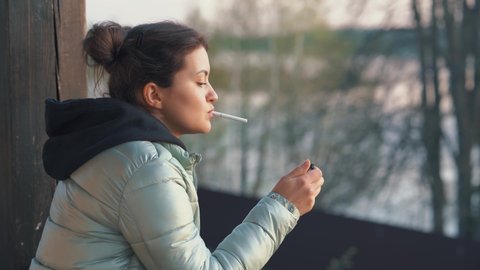 Portrait of a beautiful young woman smoking a cigarette in the evening on the veranda. Close-up of a thoughtful face of a girl looking into the distance in the open air.