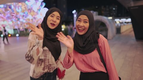 Attractive asian Muslim girls looking at camera with positive smiley face and waving hands, female tourists walk around mall with new purchases in their hands, friendship relation, relaxing activity