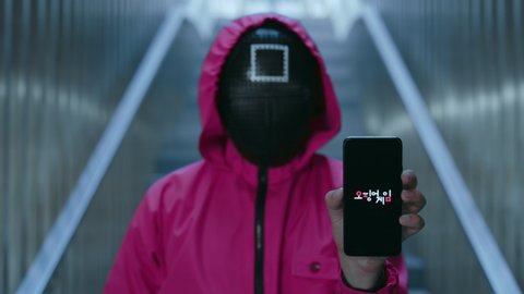NEW YORK, NY, USA - November 10, 2021: Unrecognizable man in red overalls and black mask showing logo of korean serial squid game on smartphone screen. Survival drama from netflix