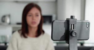 Female blogger record video vlog using smart phone seated on couch alone in living room shooting video, blurred woman silhouette. Focus on phone with tripod.