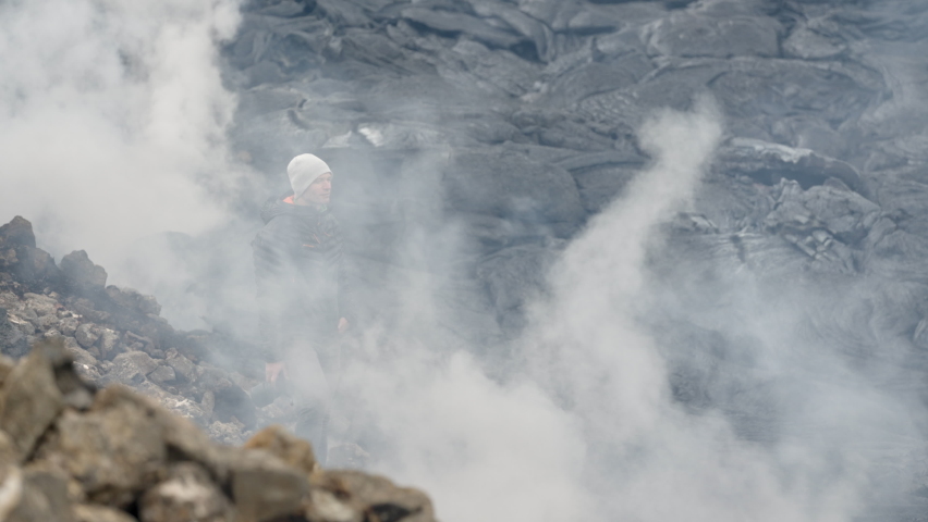 Handheld Wide Slow Motion Shot Of Man Standing With Camera Amongst Smoke Above Lava Field Of Volcanic Rock, Iceland
