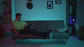 Handsome Isolated Asian Man Laying on a Dark Grey Sofa in His Moody Bedroom Using a Black Tablet for Scrolling on Social Media and Watching Videos, Relaxing After Remote Working From Home Office.