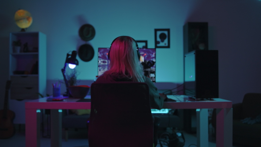 Beautiful Isolated Caucasian Gamer Girl Sitting in Her Moody Bedroom In Front of a Computer Screen Playing Games Before Turning Around To Look Directly at the Camera, Smiling, Wearing a Comfy Hoodie. Royalty-Free Stock Footage #1082798242
