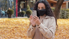 Beautiful girl in medical mask stand in autumn park hispanic young woman making video call outdoors uses app on telephone female looking at phone screen lady typing on device protection against covid