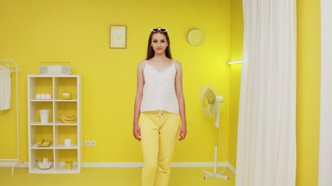 Portrait of young confident woman in white top and yellow trousers, beautiful model, standing in bright yellow interior, posing on camera and showing thumbs-up gesture, Slow motion.