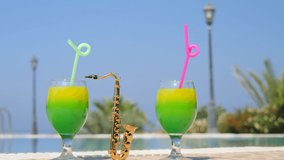 Two bright beautiful yellow-green cocktails with straws glasses, mini alt golden saxophone stand on side of pool, against background of blue water. Summer party, poolside relaxation, vacation video.