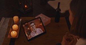 View from shoulder of young woman having video call on digital tablet with her lovely boyfriend. Happy caucasian couple chatting, smiling and drinking wine during online dating.