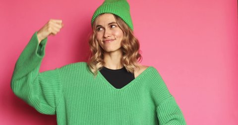 Attractive blonde woman in green knitted hat and sweater isolated on pink background. Feminism and women power. Do it! Self assured proud woman shows muscles in hands. I am strong and independent!