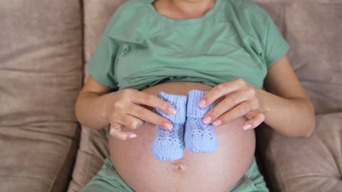 Pregnant, beautiful woman sitting on the sofa with little blue baby booties. Woman moves the tiny socks back and forth on her big bare belly.