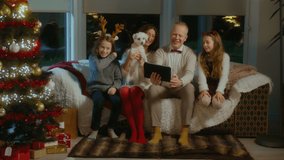 Beautiful Caucasian Family Having a Video Call With Grandma, Using a Black Tablet, Showing Off Their New White Dog. Sitting Together on a Comfortable Sofa in a Festive Decorated Living Room.