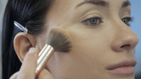 A young woman is preparing for a photo session. Close-up of a makeup artist's hand applying powder to the model's face with a brush. Beauty industry