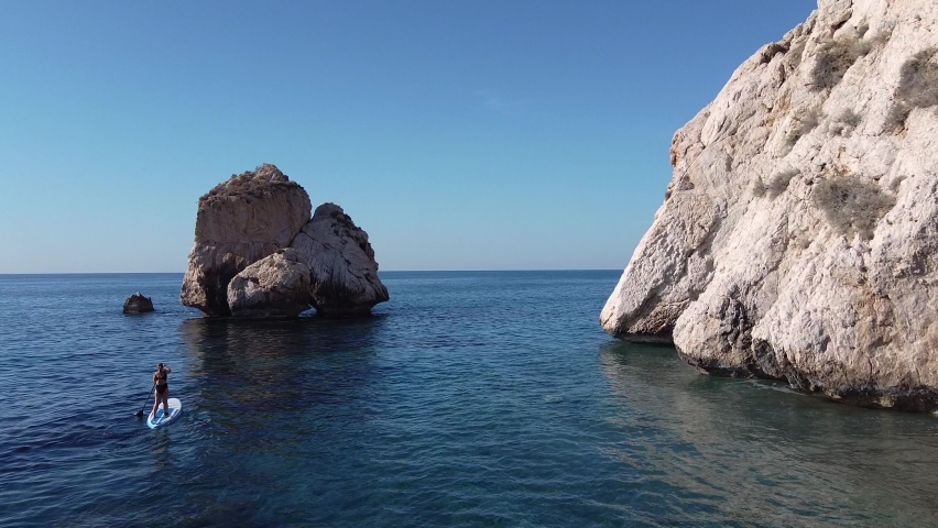 Scenic view of a woman paddleboarding by large rocks in the Mediterranean Sea.