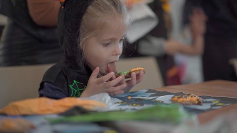 Little girl eat icing just making handmade Halloween cookies sitting on the table dressed Halloween carnival costume with horns . Halloween party in big family. High quality 4k footage