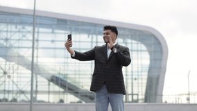 People, lifestyle, travel, tourism and modern technology. Handsome happy Indian businessman making video call on smartphone to his friend and showing hello gesture against modern airport background