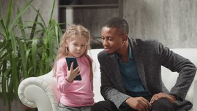 Cute blonde little girl shows favorite cartoons via mobile phone to positive African-American man sitting on large sofa in living room slow motion