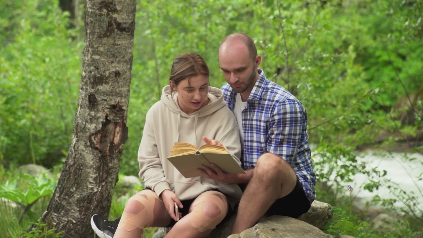 A man and a woman are sitting on a rock. A man is reading a book with a friend. A boy and a girl have a date. In the forest in the open air