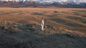 Video of kungfu master in a white sports uniform training on the hill. Man perform wushu forms in nature on background of snowy mountains at sunset time.