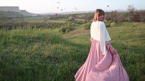 Red haired thin beautiful young girl in a pink long dress fluttering in the wind and a white jacket falling from her shoulders runs down the slope to the colorful balloons. 4k cinematic video
