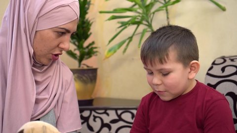 A Muslim mother in a hijab teaches her son to read a book at home