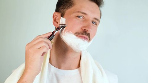 Handsome young man with towel on shoulders and moisturizing foam on face shaving with modern sharp razor and looking at camera indoors. Shaving, men's beauty and daily skin care concept.
