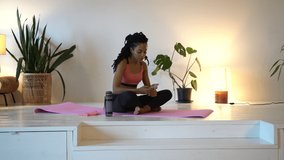 Attractive young lady in sports wear sitting on yoga mat with crossed legs and using smartphone after trainings. Relaxing woman having break at living room
