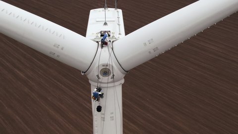Service engineer and coworker high-altitude installer with safety belts go down to windmill propeller fastened by ropes above plowed field aerial view