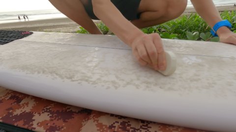Close-up of a surfer smearing wax on his surfboard against the background of the beach and the company of people playing ball. A man prepares his surfboard by rubbing it with hard wax. Slow motion.