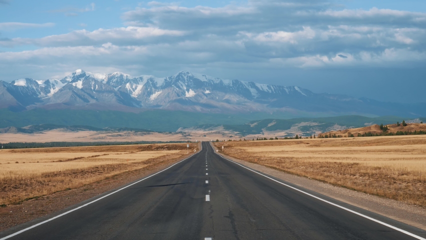 Driving on highway of black asphalt and white road markings at sunny day, road going into distance through desert prairie at autumn to snowy mountain summits on horizon in wild. Pov from moving car
