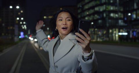 Cinematic shot of happy smiling mature asian businesswoman is making technology video call with smartphone to friends or family in busy city center after finishing work day at evening.