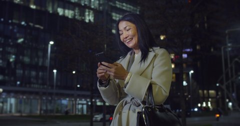 Cinematic shot of happy smiling mature asian business woman is using smartphone for work or entertainment in busy city center after finishing work day at evening.