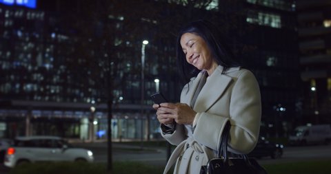 Cinematic close up shot of happy smiling mature asian business woman is using smartphone for work or entertainment in busy city center after finishing work day at evening.