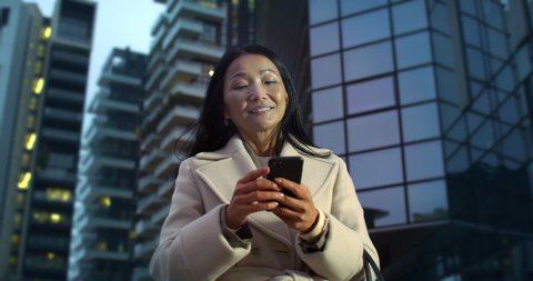 Cinematic shot of happy smiling mature asian business woman is using smartphone for work or entertainment in busy city center after finishing work day on background of skyscrapers at evening.