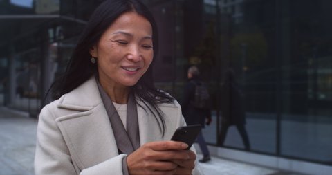 Cinematic close up shot of mature asian business woman is using smartphone for work or entertainment while walking in busy city center on way to office before to start her work day.