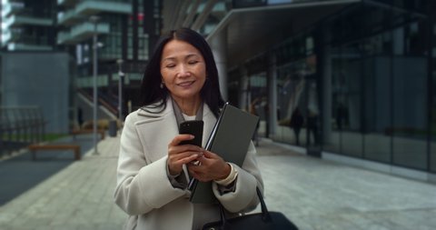 Cinematic shot of happy smiling mature asian business woman is using smartphone for work or entertainment while walking in busy city center on way to office before to start her work day.