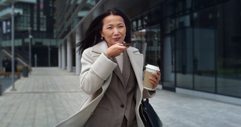Cinematic shot of happy smiling mature asian business woman is making call with smart phone and drinking morning coffee while walking in busy city center on way to office before to start her work day.