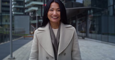 Cinematic shot of mature asian business woman is using smartphone for work and smiling in camera in busy city center before to start her work day in office.